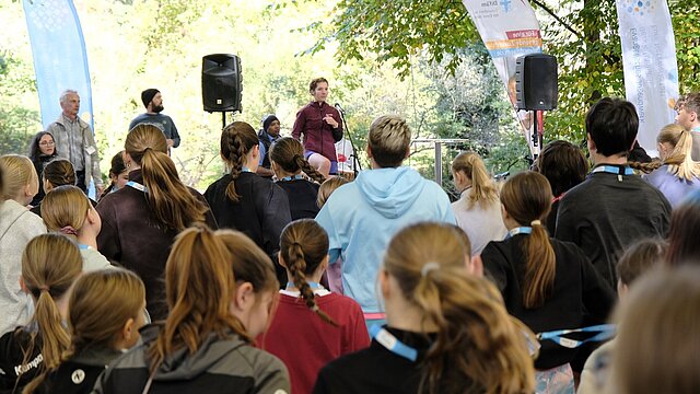 Konfirmand:innen beim Aufwärmen für den Lauf, Ansicht von hinten. Auf der Bühne steht eine junge Frau, die die Aufwärm-Übungen anleitet.