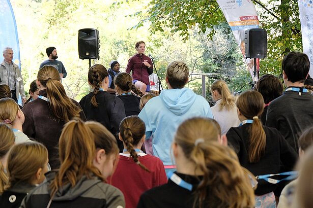 Konfirmand:innen beim Aufwärmen für den Lauf, Ansicht von hinten. Auf der Bühne steht eine junge Frau, die die Aufwärm-Übungen anleitet.