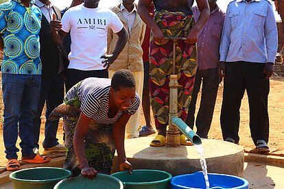 Eine Frau pumpt am Brunnen Wasser, das in mehrere Eimer sprudelt. Eine andere Frau wechselt die Eimer aus. Daneben stehen einige Männer.